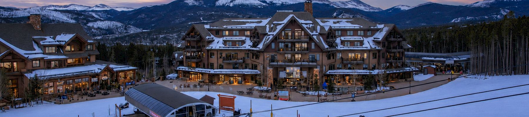 A snowy ski resort at dusk with a lodge complex, chairlift, and mountain peaks under colorful clouds.