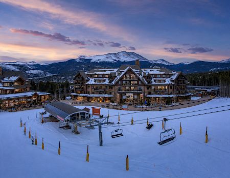 A snowy ski resort at dusk with a lodge complex, chairlift, and mountain peaks under colorful clouds.
