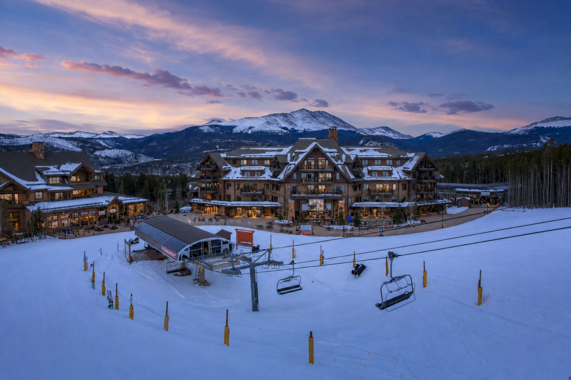 A snowy ski resort at dusk with chalet buildings, lit windows, and ski lifts below snow-capped mountains under purple clouds.