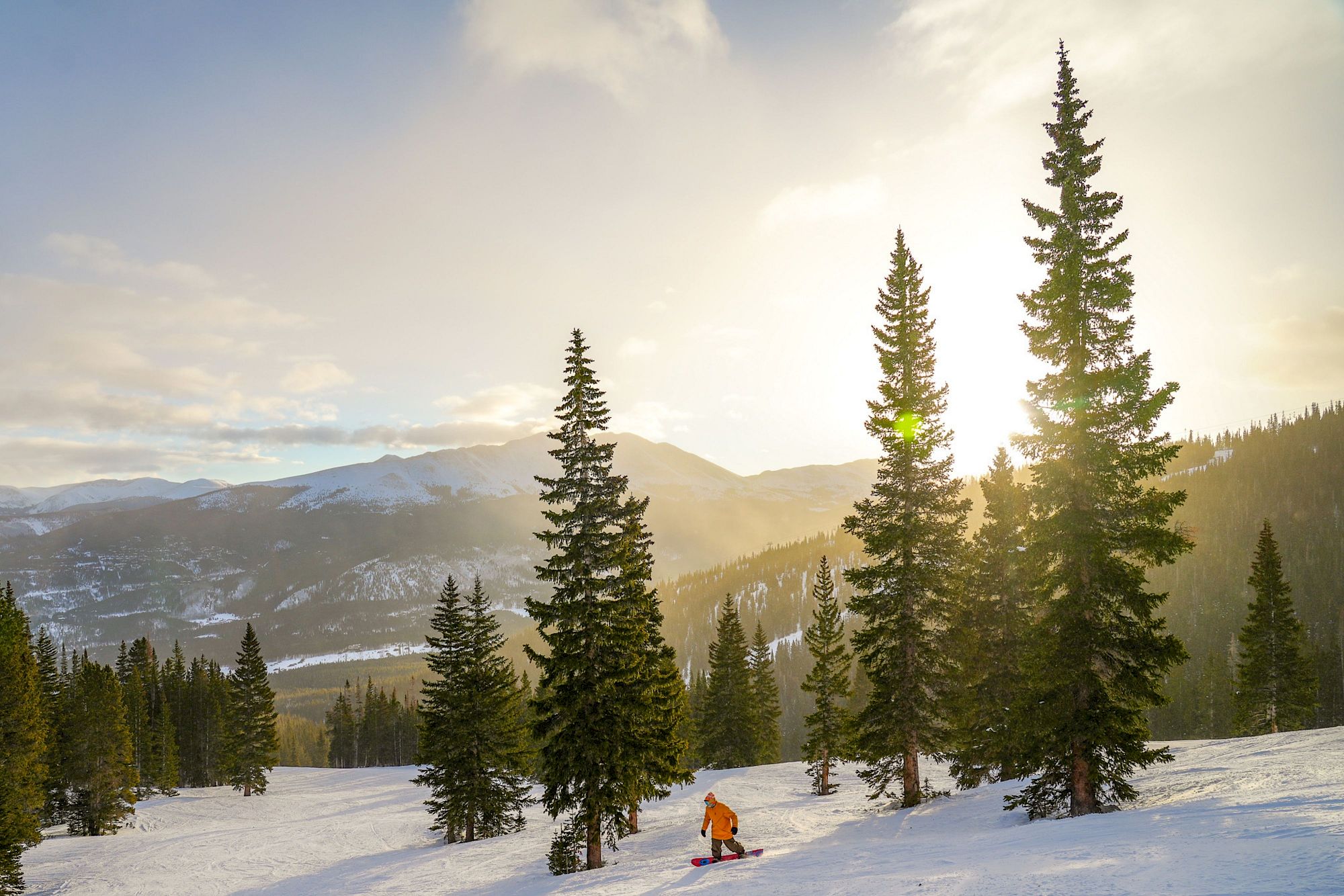 Snowy mountain valley with pine trees, golden sunrise, and a skier or snowboarder on the slope below, surrounded by distant peaks.