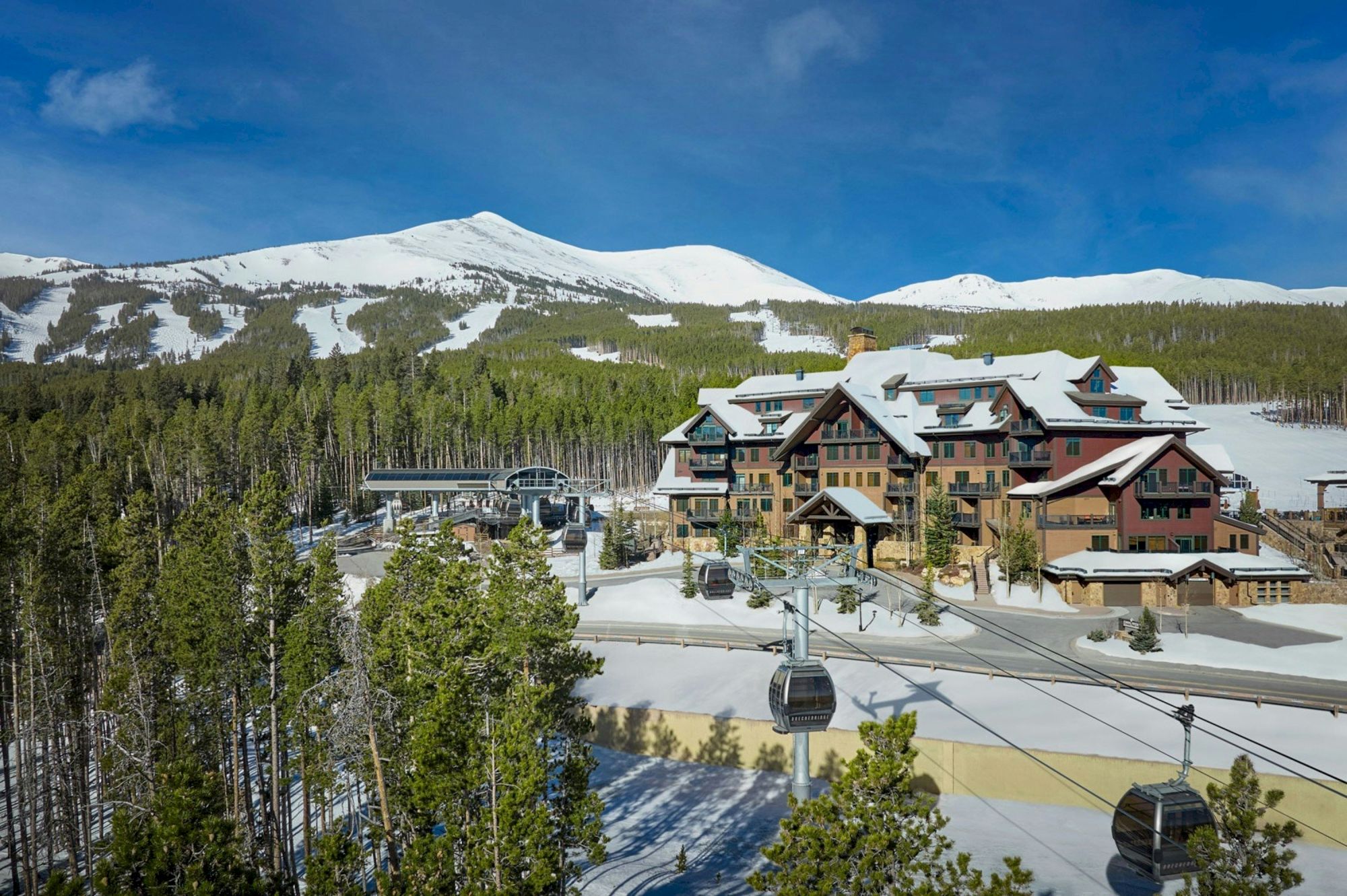 A snowy mountain ski resort with a large lodge, pine forest, ski slopes, and chairlift/gondola cables under a blue sky.