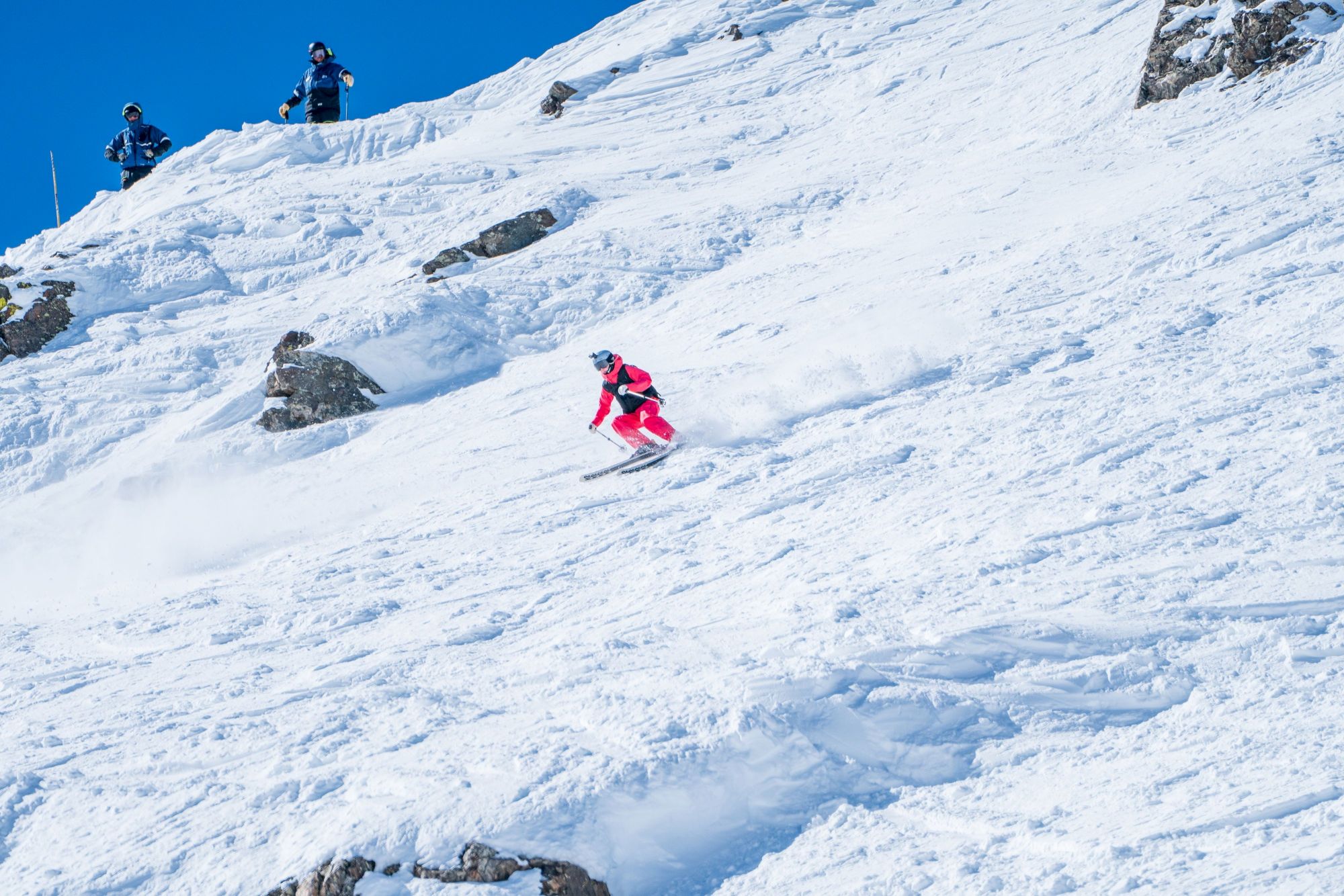Skiers in helmets carve down a steep snow-covered mountain slope under a clear blue sky.