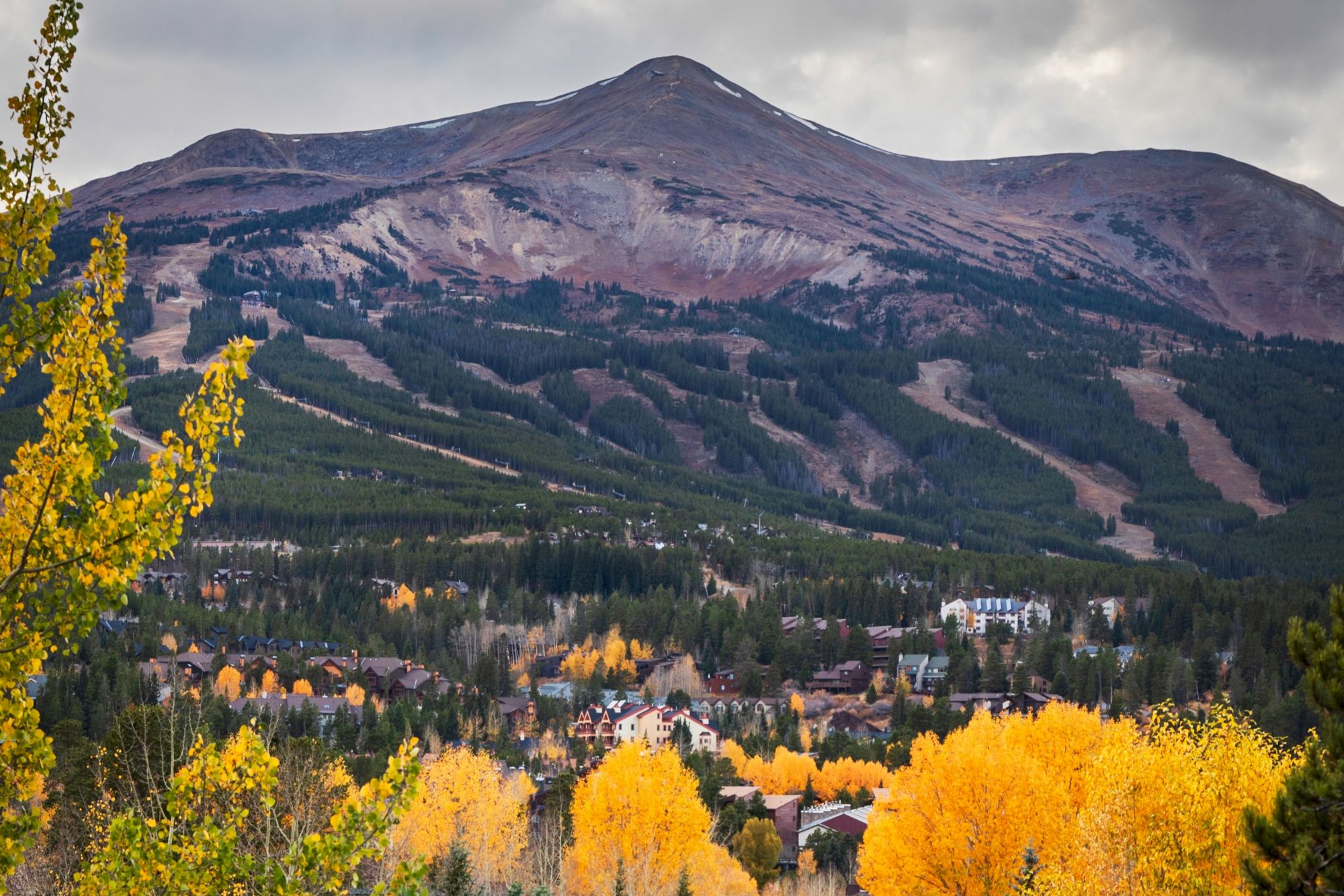 A dramatic mountain rises over a valley town, framed by bright autumn trees and cloudy skies.