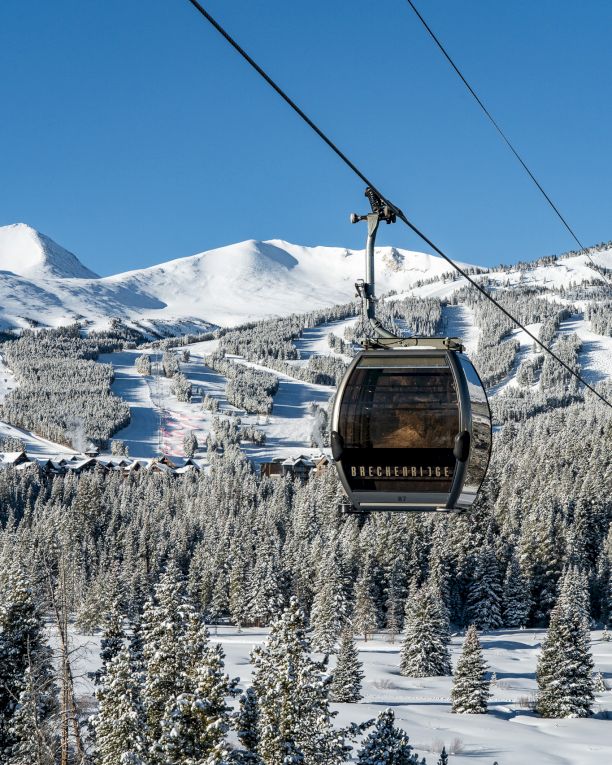 A Breckenridge gondola cable car glides over snowy pine mountains and a ski trail under a clear blue sky.