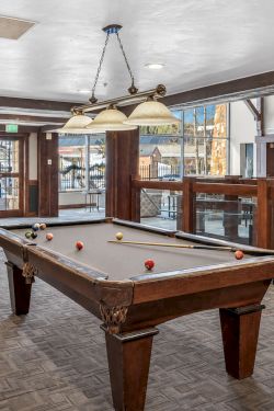 An indoor billiards room with a pool table, cues, pendant lights, and seating along a wood-paneled railing.