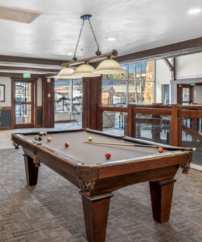 An indoor billiards room with a pool table, cues, pendant lights, and seating along a wood-paneled railing.