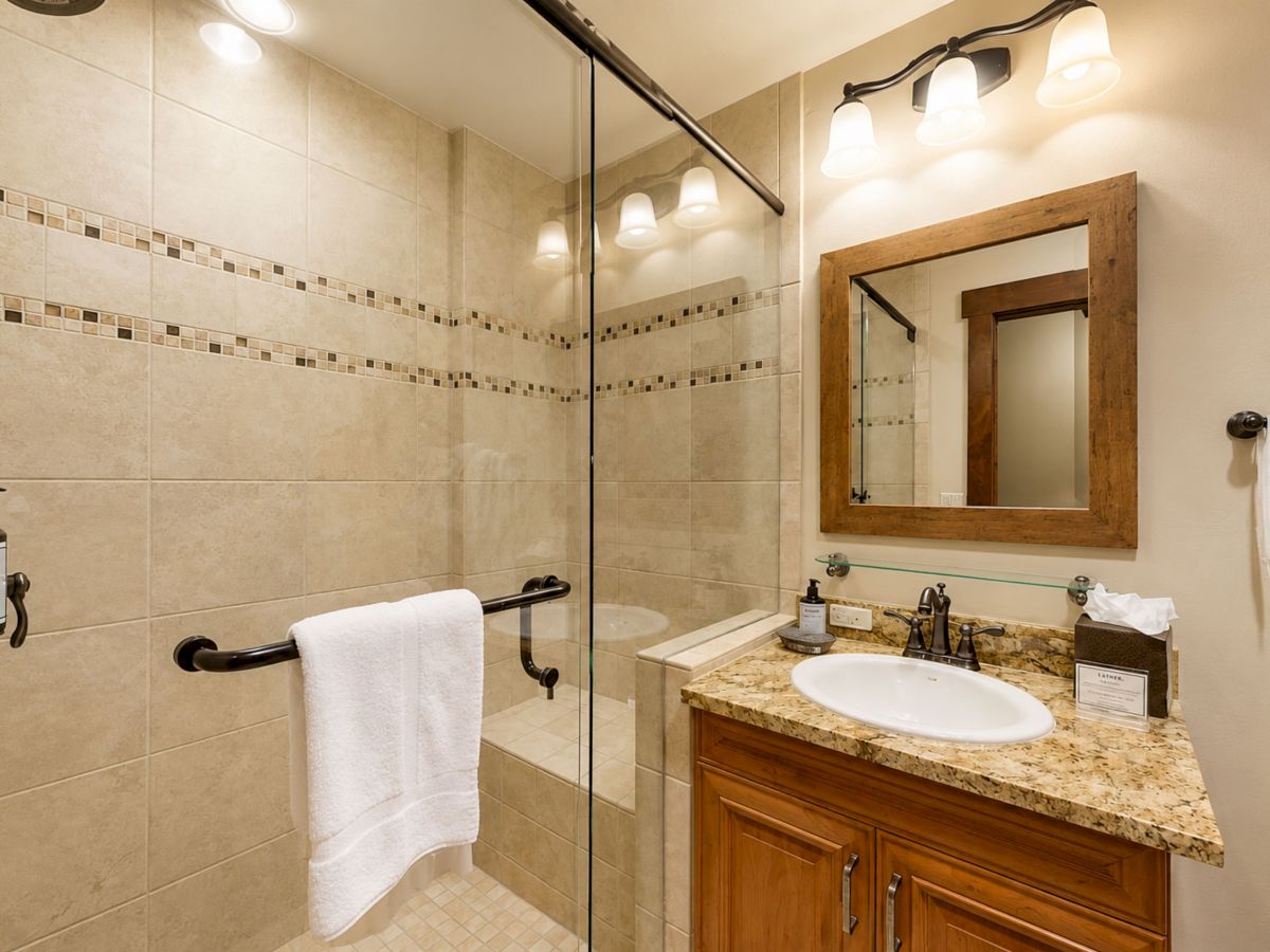 A modern bathroom with tiled shower, glass enclosure, granite vanity, sink, mirror, and bright overhead lights.