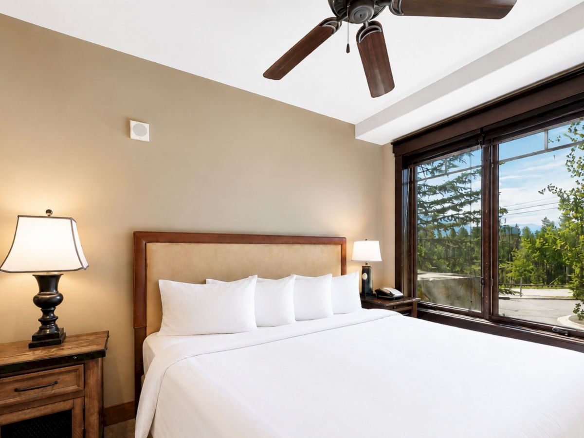 A bright hotel bedroom with a bed, bedside lamps, a ceiling fan, and large sliding windows showing trees outside.