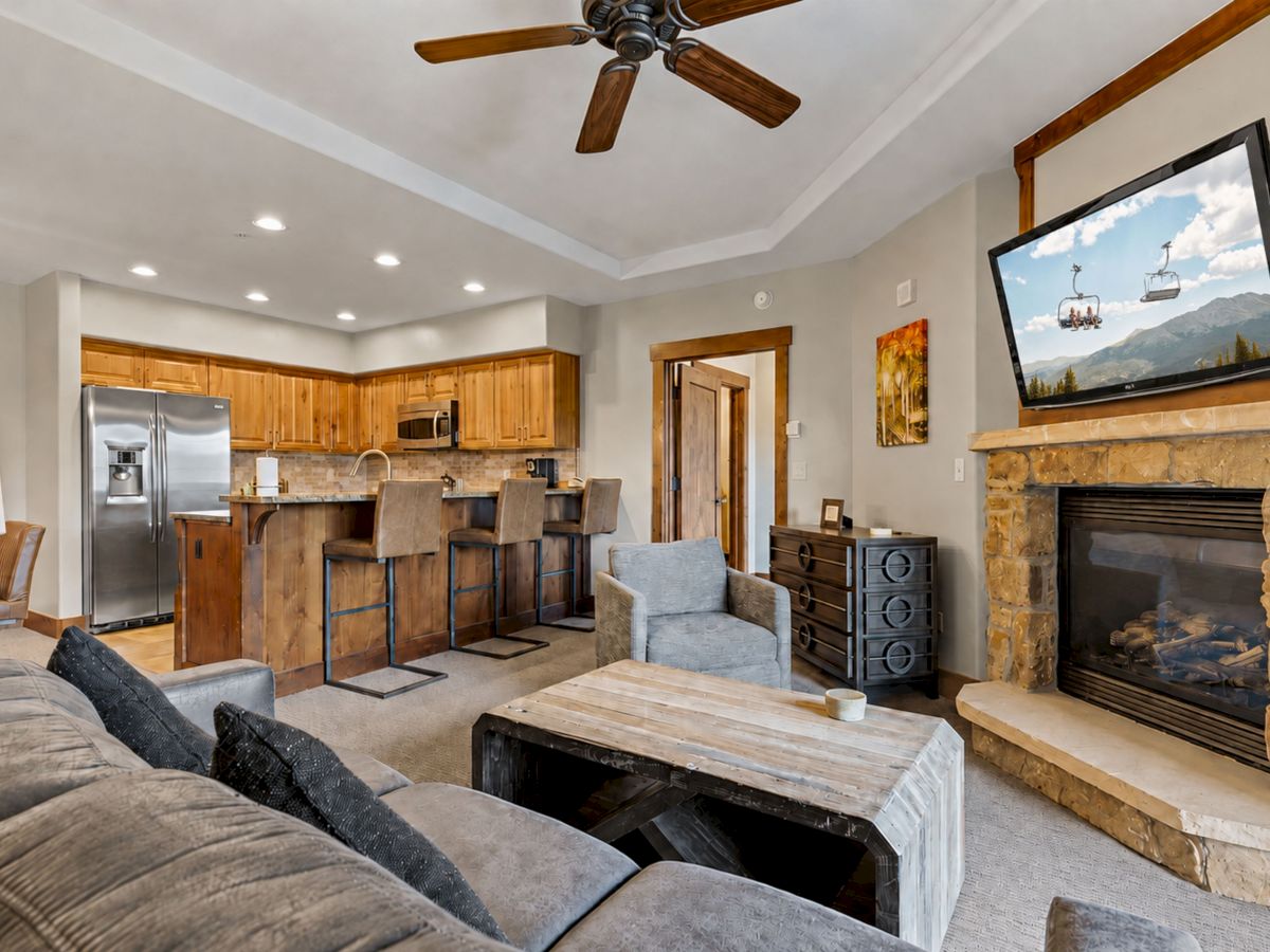 Open-plan living room with wood kitchen cabinets, bar stools, fireplace, TV, and a ceiling fan.