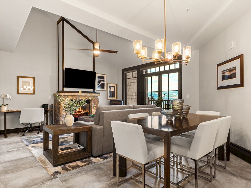 Bright modern living/dining room with gray seating, a stone fireplace, chandelier, and large windows showing an ocean view.