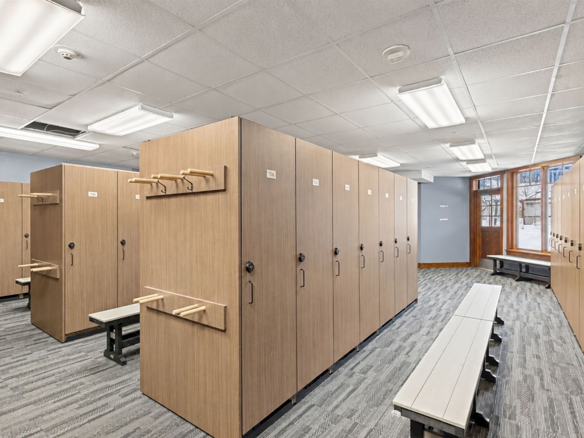 A bright locker room with rows of wooden lockers, coat racks, and benches beneath ceiling fluorescent lights.
