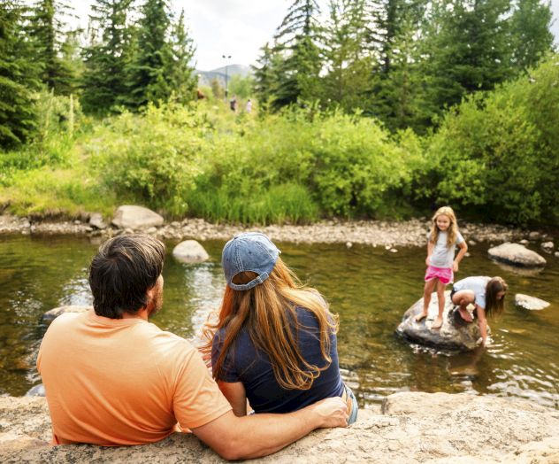 A family relaxes by a clear river while kids play in the shallow water, surrounded by lush green trees.