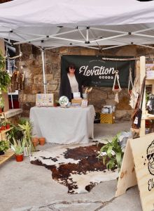 An outdoor craft market booth for &ldquo;Clevation Goods&rdquo; under a canopy, with jewelry, hanging items, and potted plants.