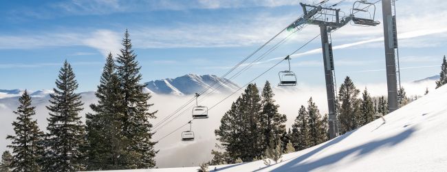 A ski resort cable lift carries empty chairs above snowy slopes, pine trees, and misty mountain peaks under a blue sky.