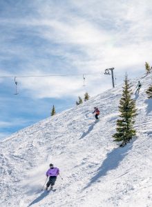 Skiers carve a snowy mountainside while a chairlift hangs overhead under a bright blue sky.