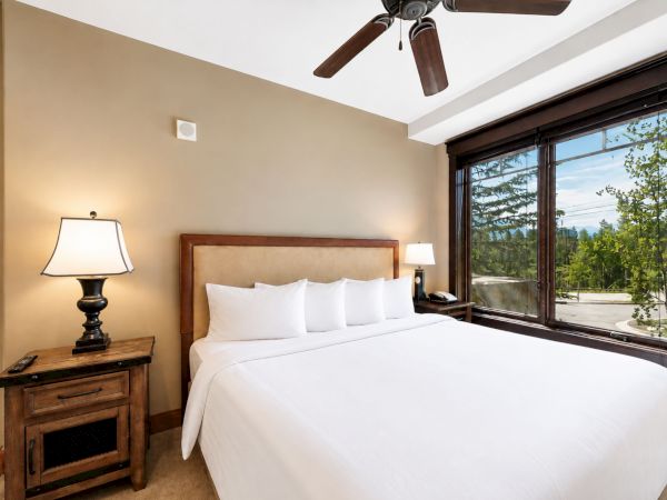 A cozy bedroom with a bed, nightstands with lamps, a ceiling fan, and large sliding windows showing greenery outside.