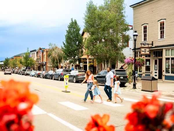 Downtown street scene with cars and pedestrians walking past shops and trees, flowers blooming along the sidewalk.