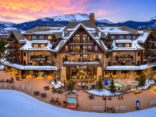 Snowy mountain resort lodge with warm lights, a courtyard, and a small tower behind it at sunset.