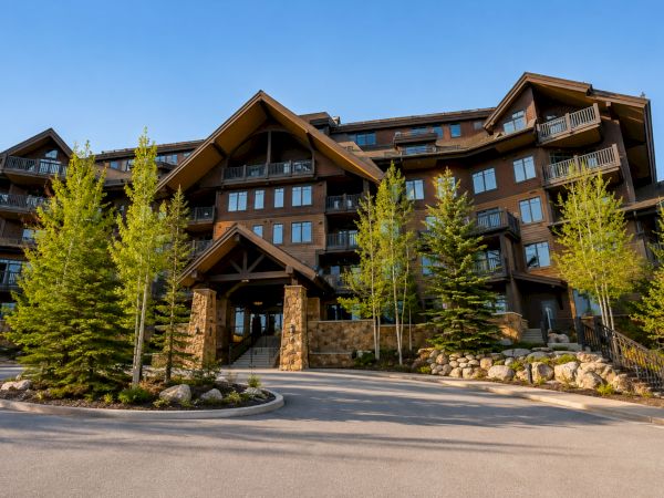 A multi-story apartment building with balconies, stone accents, and landscaping with young trees under a clear blue sky.