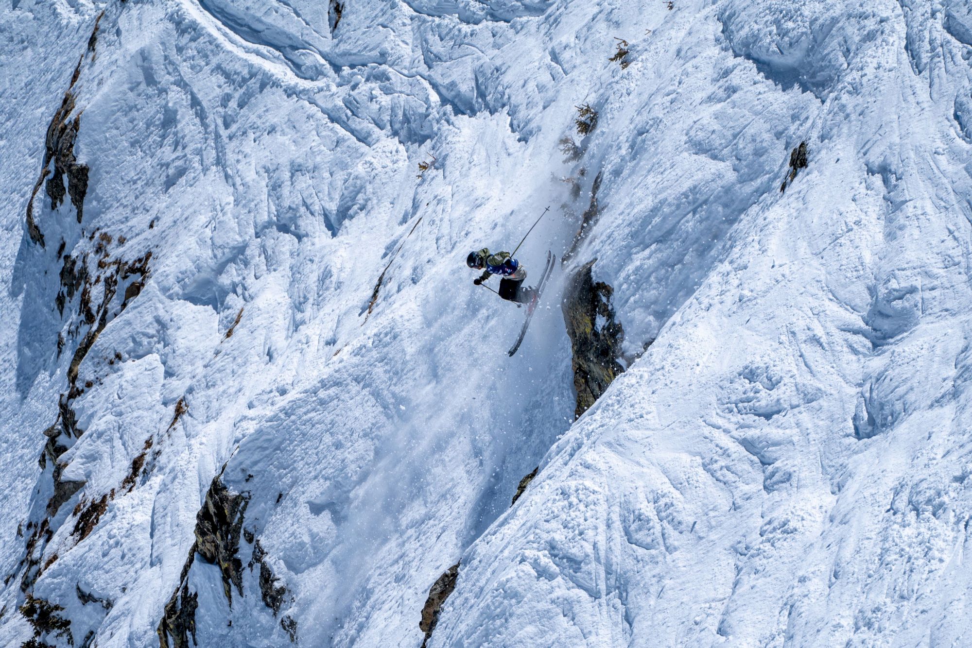 A skier in protective gear carves through heavy powder on a steep alpine slope as chunks of snow fly around.